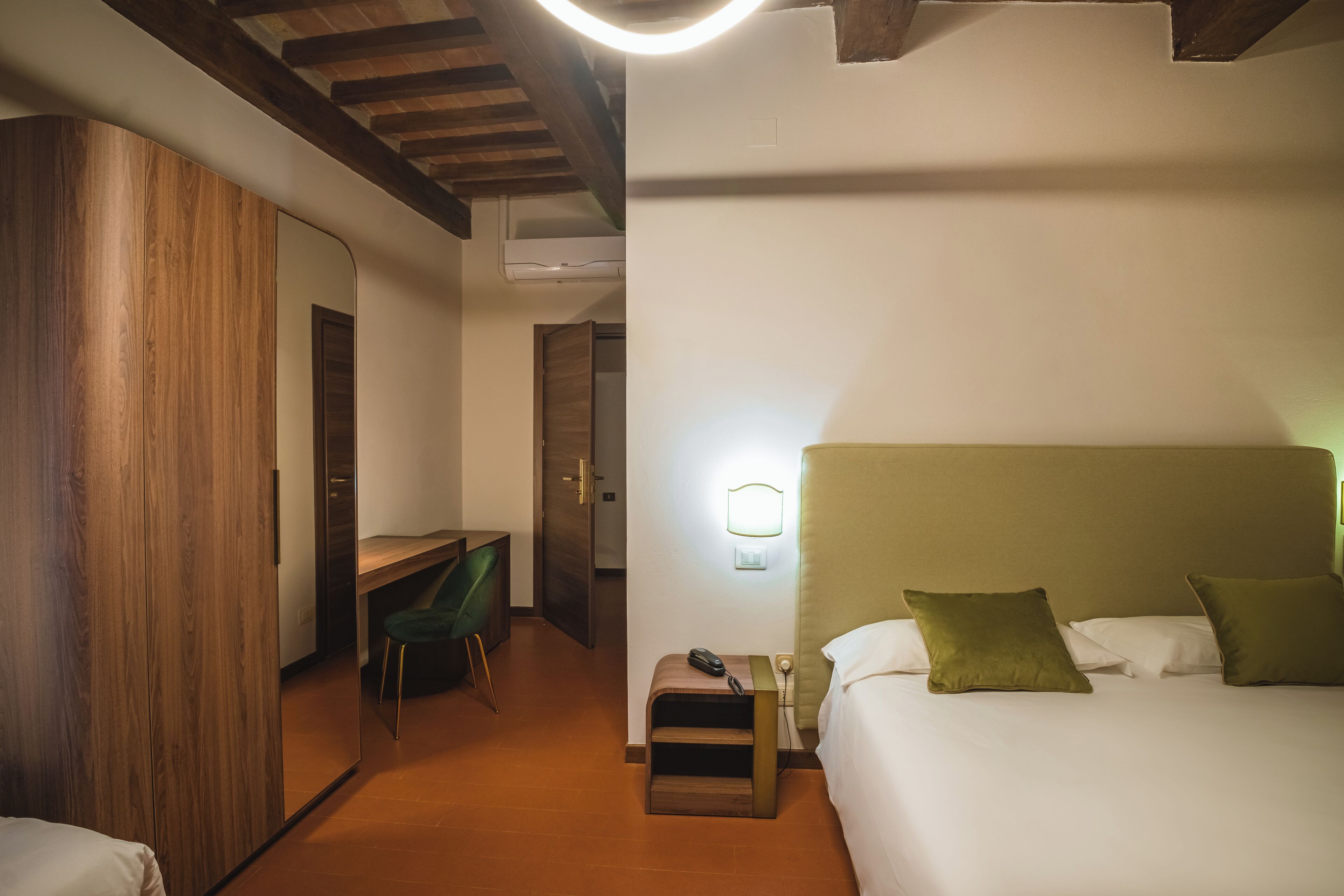 A hotel room with a wooden wardrobe, a desk with a chair, a bedside table with a lamp, and a bed with pillows.