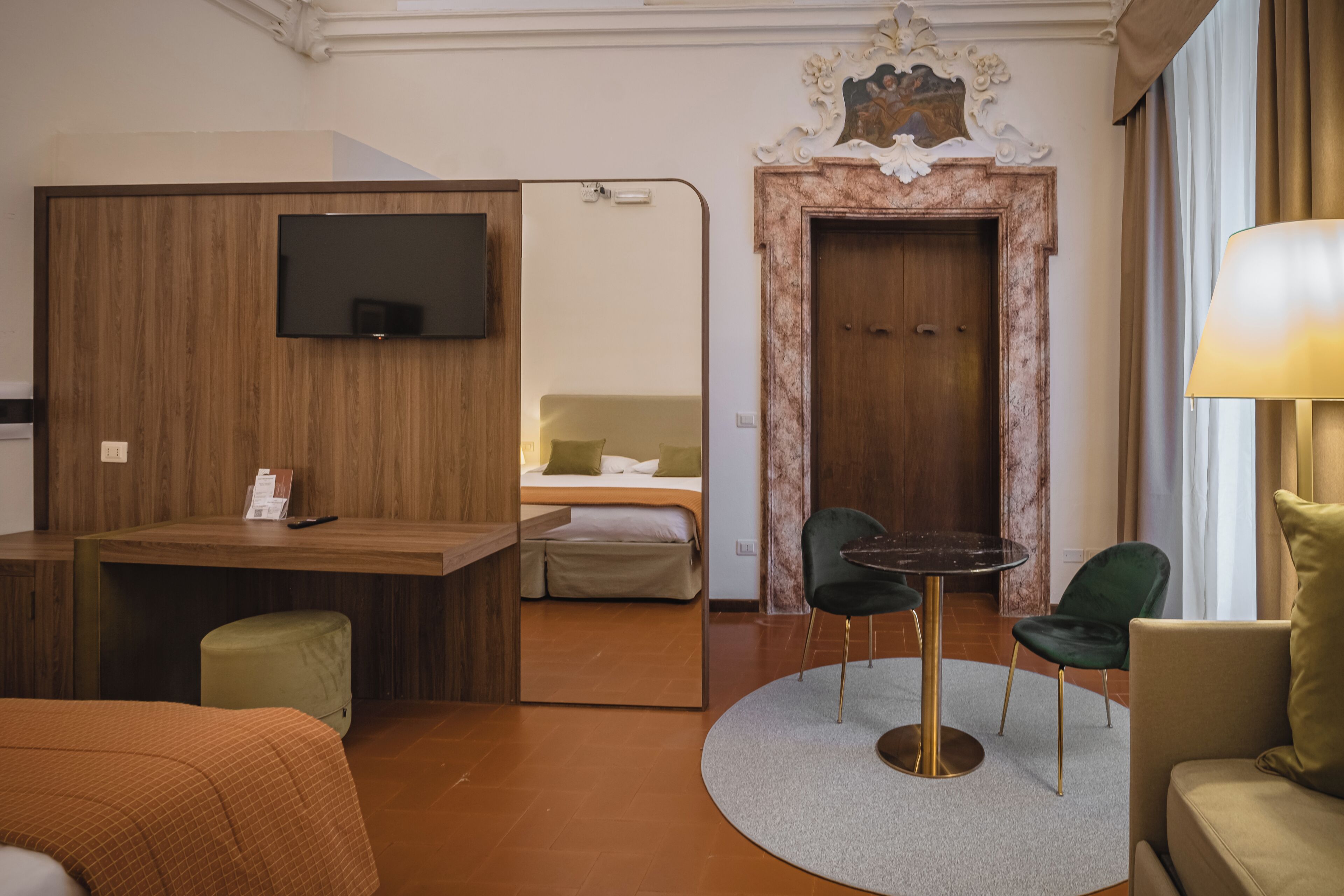 A hotel room with a wooden desk, a television, a bed with a green chair, and a round table with chairs.
