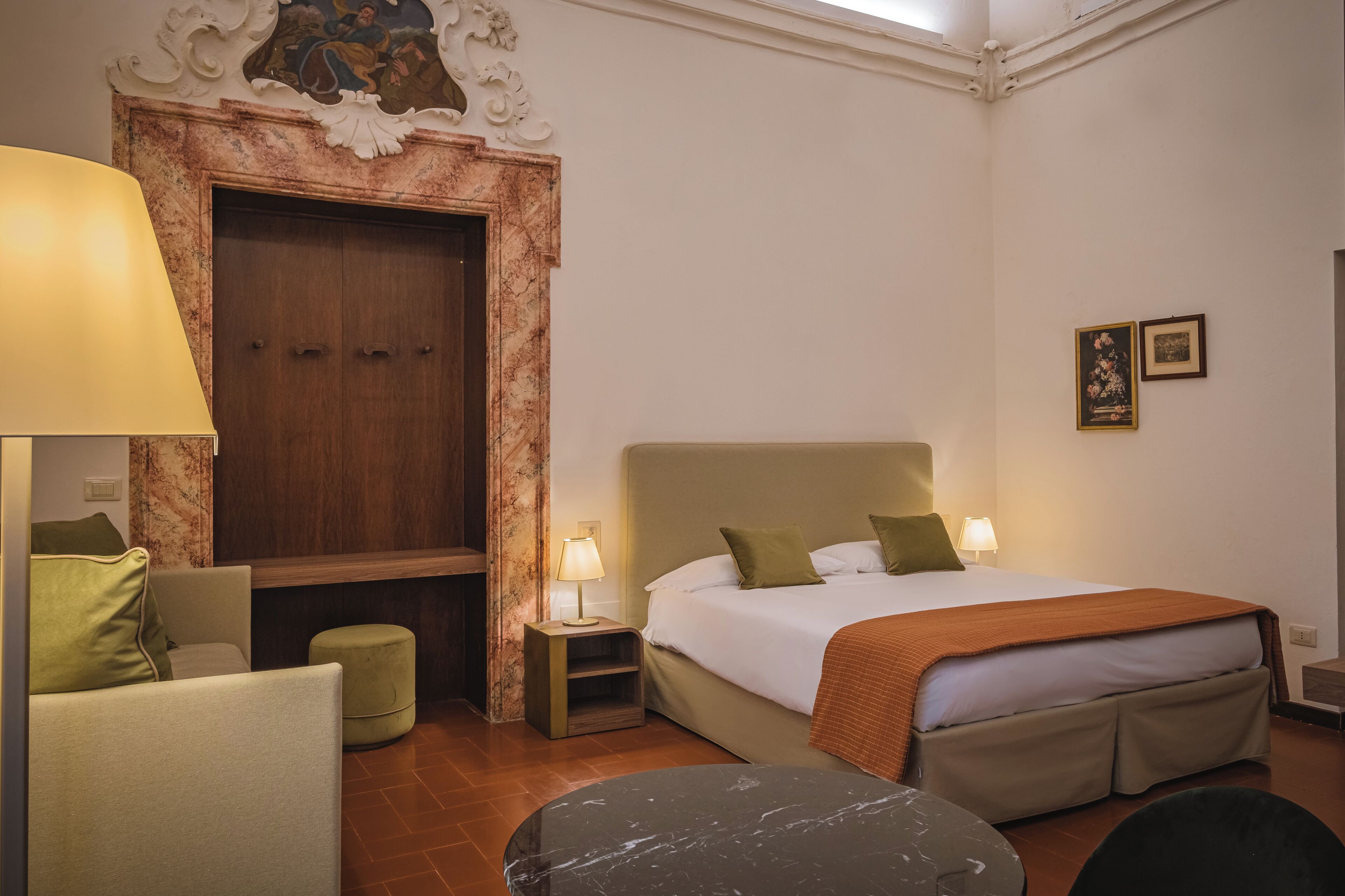 Una habitación de hotel con una cama grande, dos lámparas de noche, una mesita de mármol, un sofá y un armario de madera.