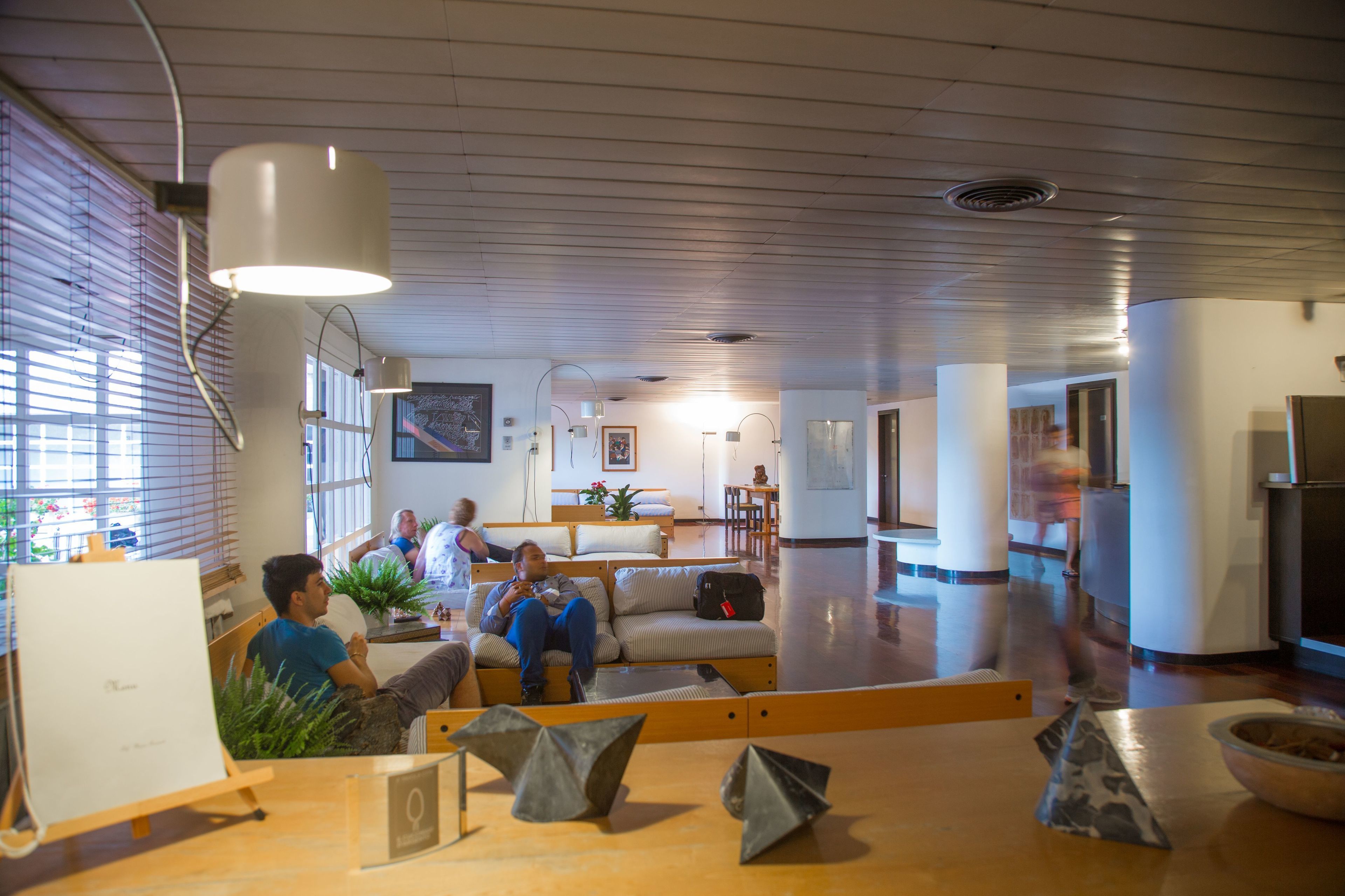 A spacious lounge area with a long wooden table, several couches, and a large window with blinds.
