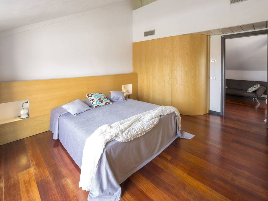 Un dormitorio moderno con cabecera de madera, una cama grande con colcha gris y una manta blanca. Se observa una mesita de noche con lámpara y un pequeño adorno.