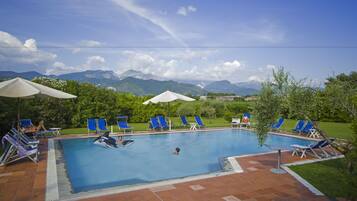 Ein Swimmingpool mit blauem Wasser, umgeben von blauen Liegestühlen und weißen Sonnenschirmen, vor einer Berg- und Grünlandschaft.