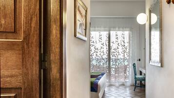 Um quarto com porta de madeira, uma cama com colcha colorida, uma cadeira, uma janela com cortinas translúcidas e um espelho na parede.