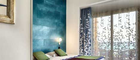 Une chambre à coucher avec un lit, des oreillers, une table de chevet, une lampe fixée au mur et une fenêtre avec des rideaux.