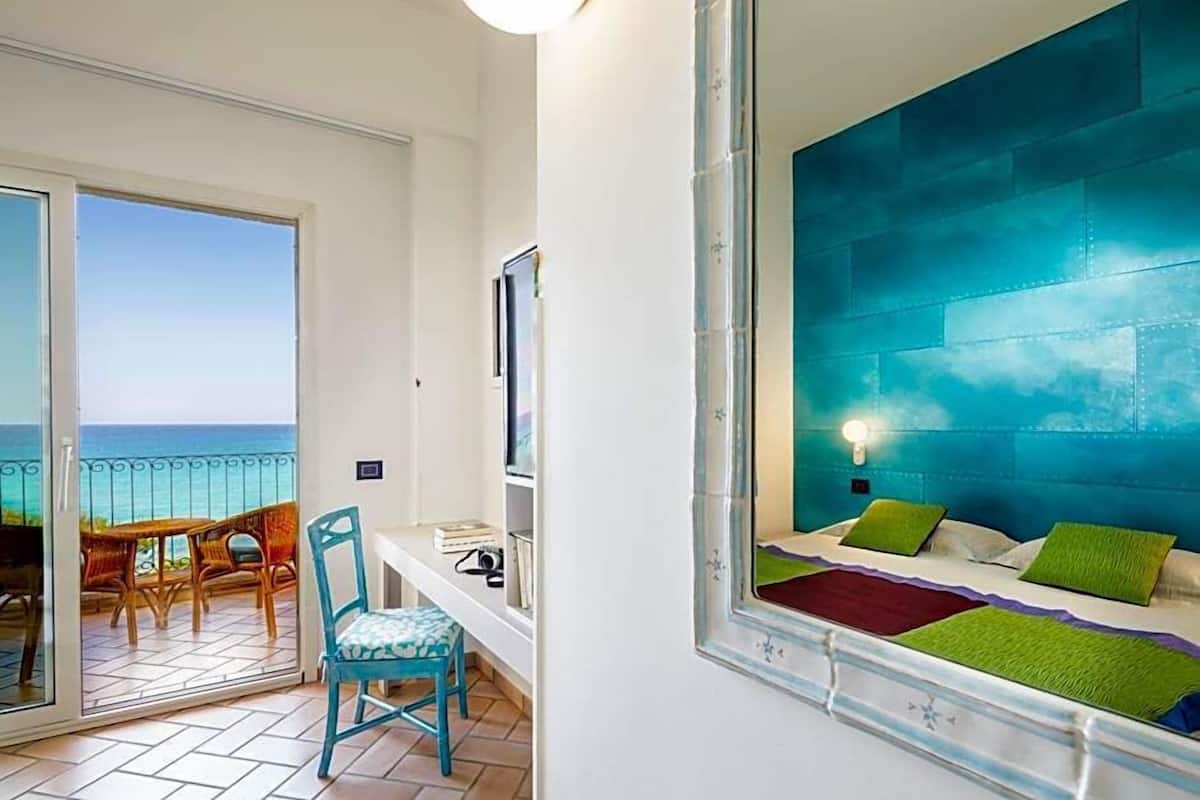 A room with a bed, a chair, a desk, a TV, and a balcony with a view of the sea.