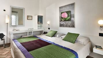 Habitación de hotel con cama, escritorio, silla, espejo, un cuadro y una mesita de noche con lámpara.