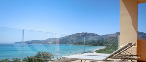 A balcony with a glass railing overlooking a beach and mountains.