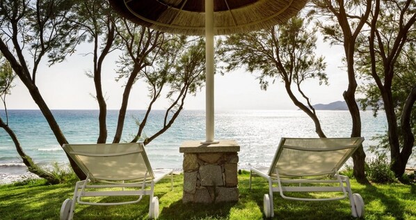 Two white lounge chairs under a thatched umbrella, positioned on a stone pedestal by a seaside view. - Hotel Stella Maris (Villasimius)