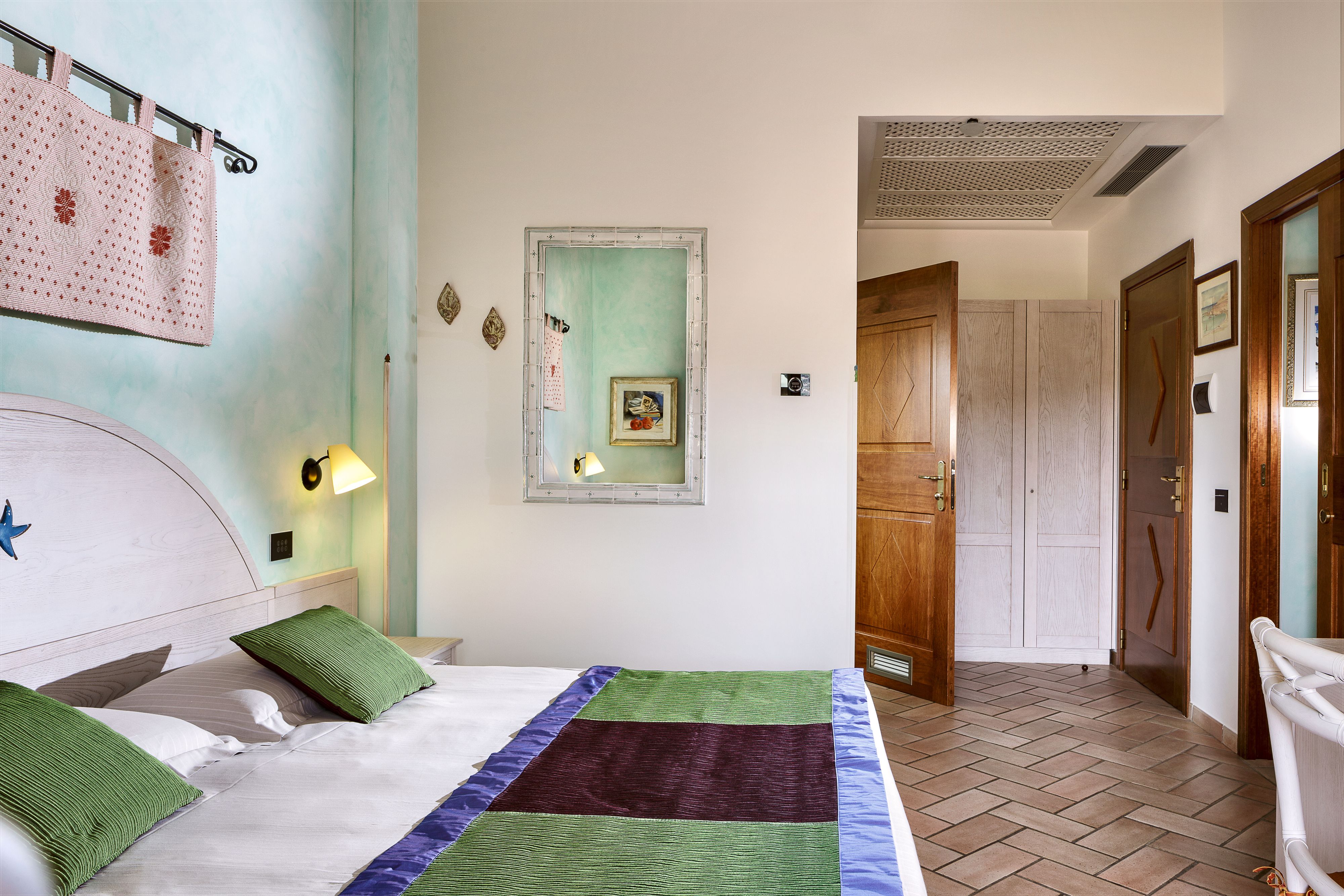 A hotel room with a bed, a nightstand, a wall-mounted lamp, a framed picture, and a wooden door.