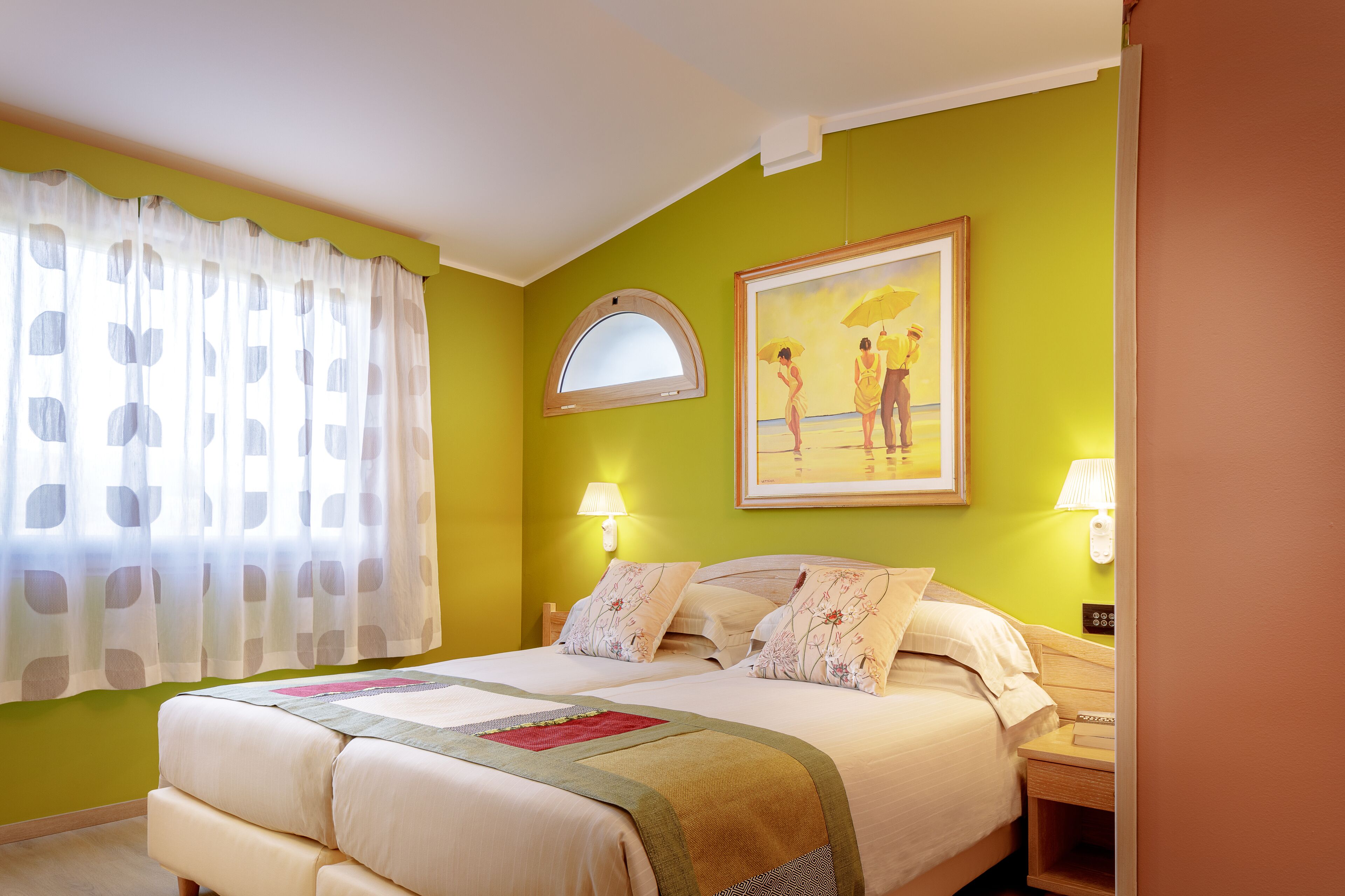 A hotel room with a bed, two bedside tables, a window with curtains, a painting on the wall, and a lamp on each bedside table.