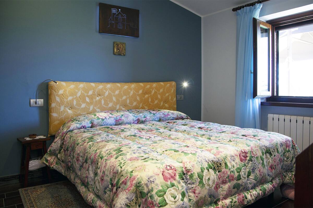A bedroom with a floral bedspread, a nightstand, a wall-mounted picture, and a window with curtains.