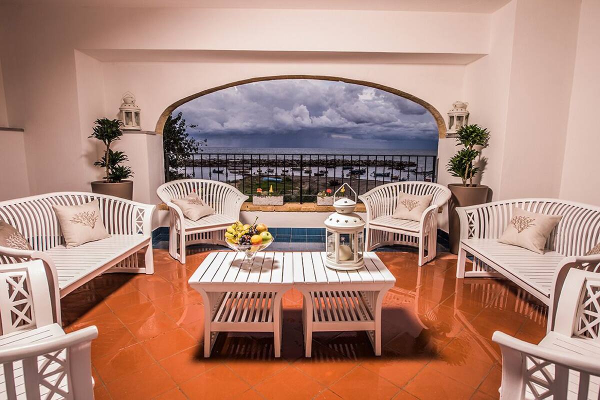 A terrace with white wicker furniture, a table with fruit, and a view of the sea through an archway.