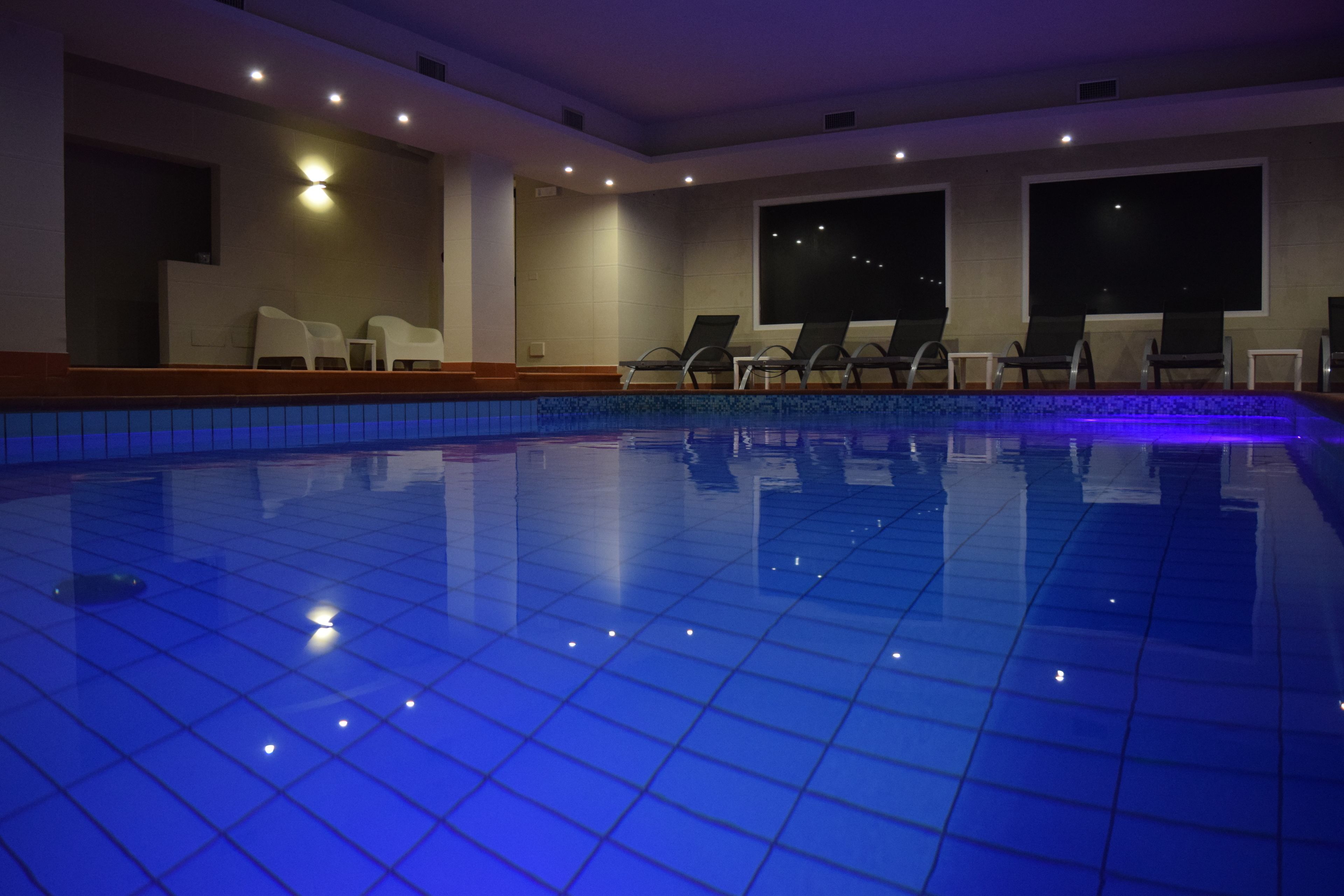 En indendørs swimmingpool med blå flisegulv, liggestole og udsigt til natten gennem store vinduer.