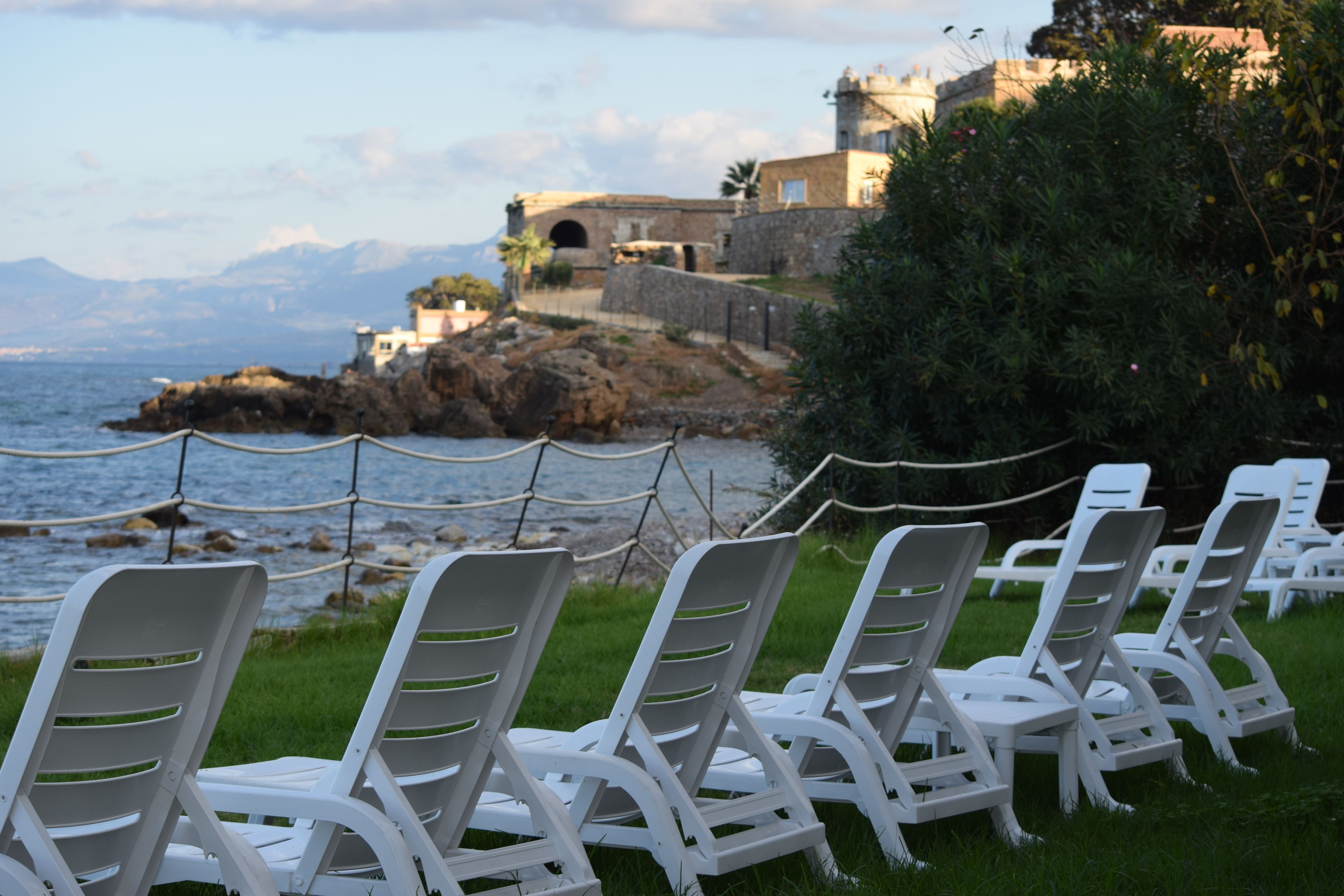 White plastic chairs arranged on grass near a rocky coastline with a building in the background.