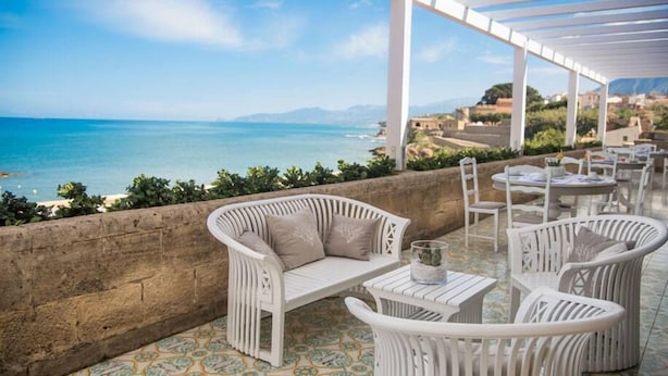 A terrace with white wicker furniture, a view of the sea, and a mountainous backdrop. - Hotel Tonnara (Trabia)