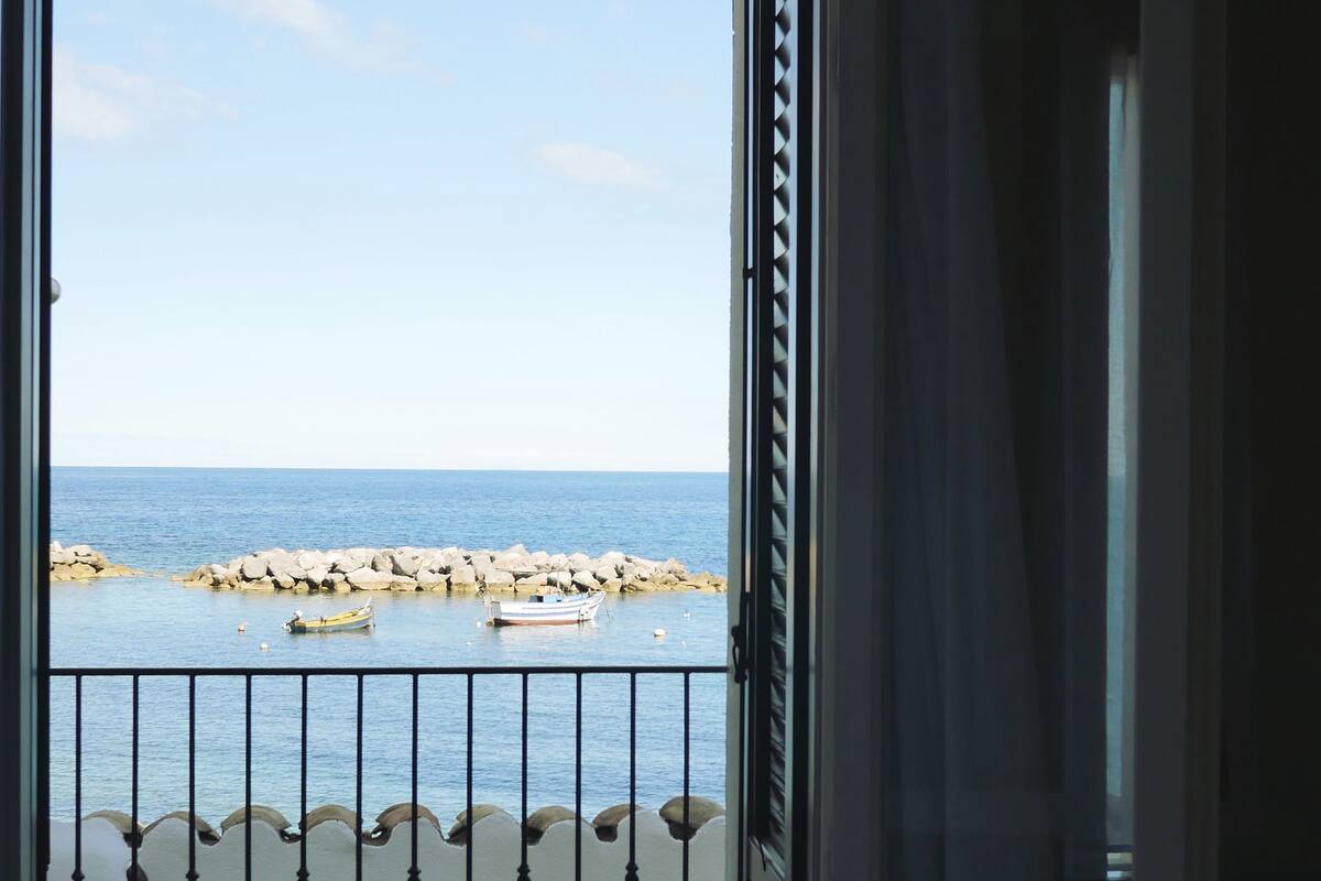 A room with a bed and a balcony with a view of the sea and rocks.