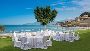 An outdoor wedding setup with tables and chairs arranged on a grassy area, overlooking a body of water and a historic building. - Hotel Tonnara (Trabia)