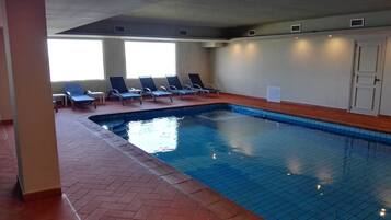 En indendørs swimmingpool med flisebeklædte vægge og et lagt gulv. Liggestole står langs poolkanten.