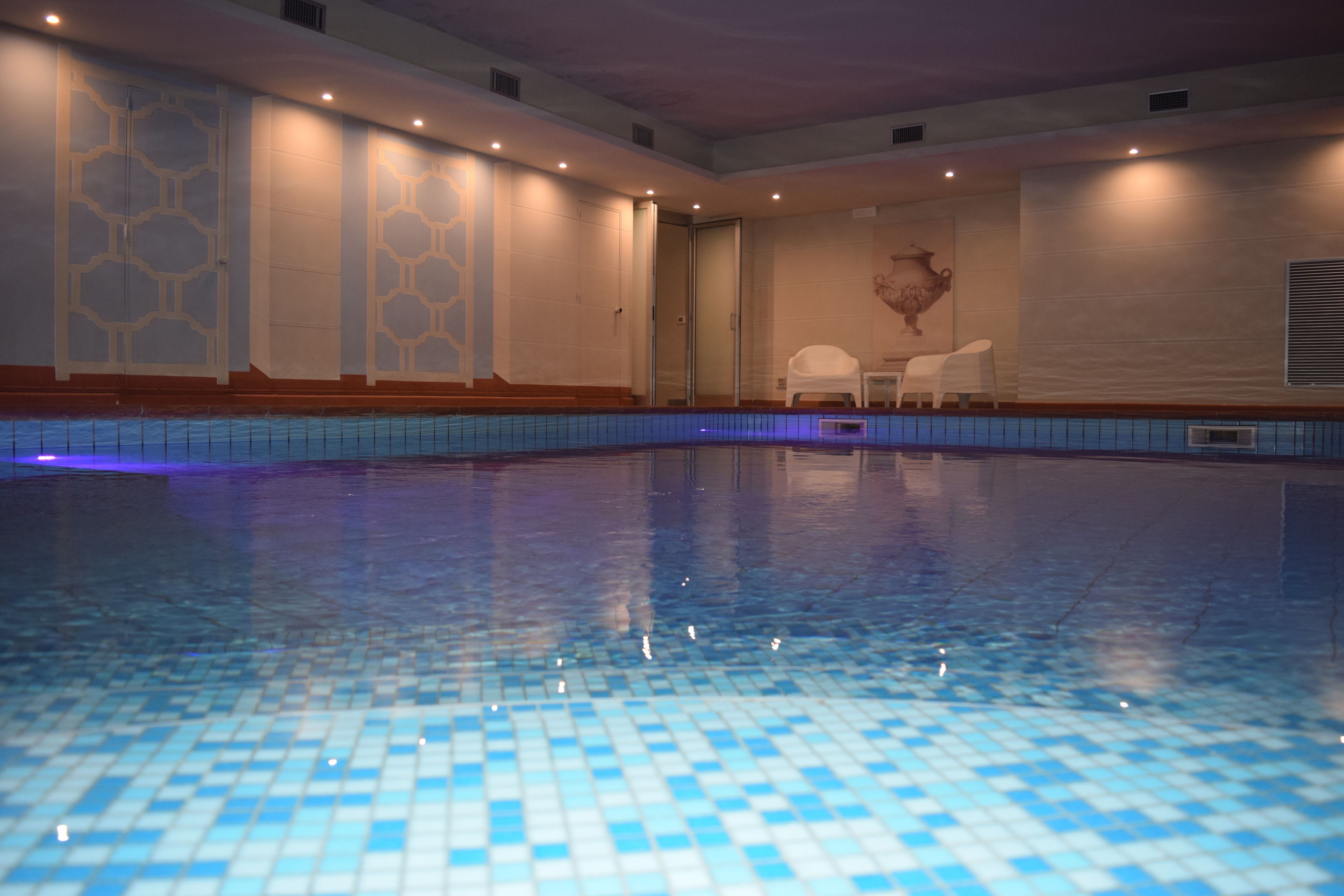 A large indoor swimming pool with blue mosaic tiles, a white wall with a geometric pattern, and a white chair near the wall.