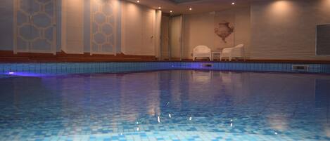 A large indoor swimming pool with blue mosaic tiles, a white wall with a geometric pattern, and a white chair near the wall.