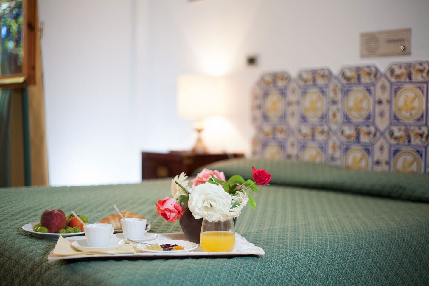 Una bandeja de desayuno ordenada sobre una cama con edredón verde, un florero y una vista de una pared estampada y una lámpara al fondo.