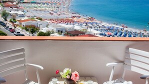 Un balcon avec des chaises blanches et une table, donnant sur une plage parsemée de parasols et fréquentée par des gens.