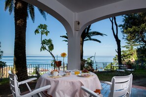 Une table dressée pour le petit-déjeuner, avec vue sur la mer, entourée de chaises blanches et recouverte d’une nappe blanche.