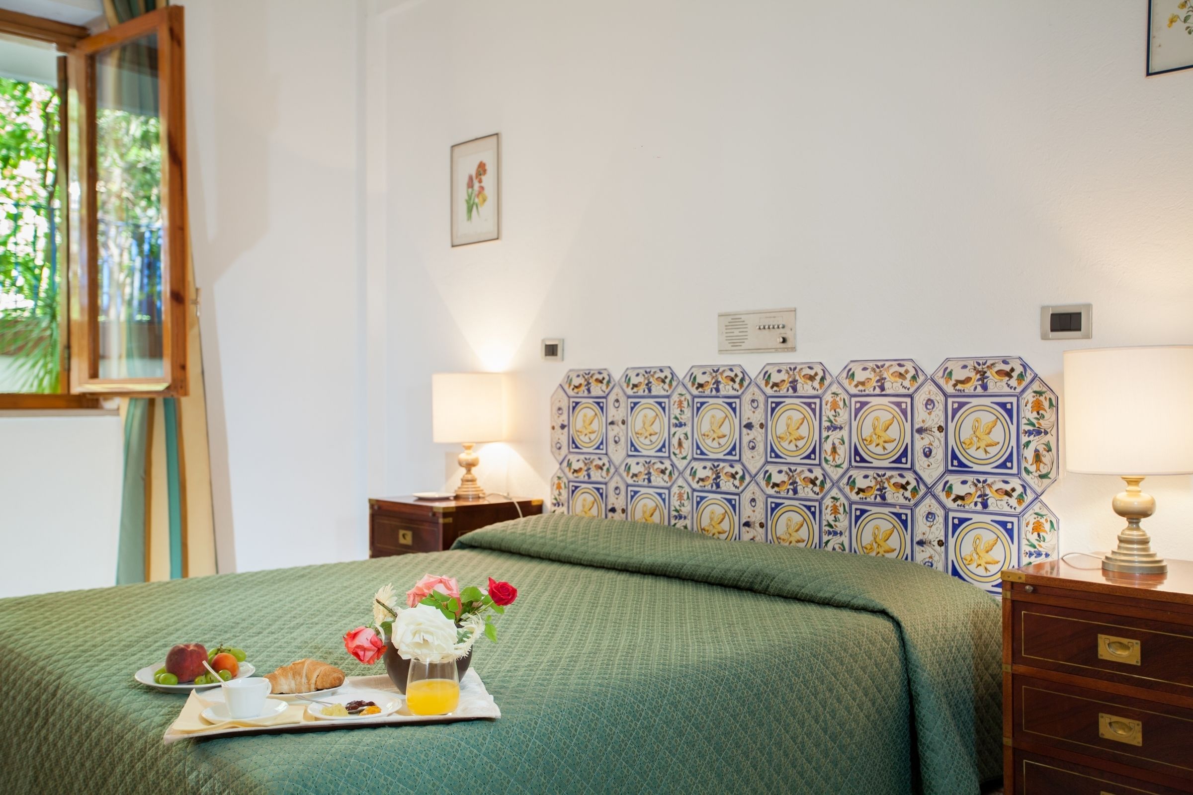 Una cama bien tendida con una colcha verde, una bandeja con alimentos para el desayuno y una pared decorada con baldosas hexagonales.