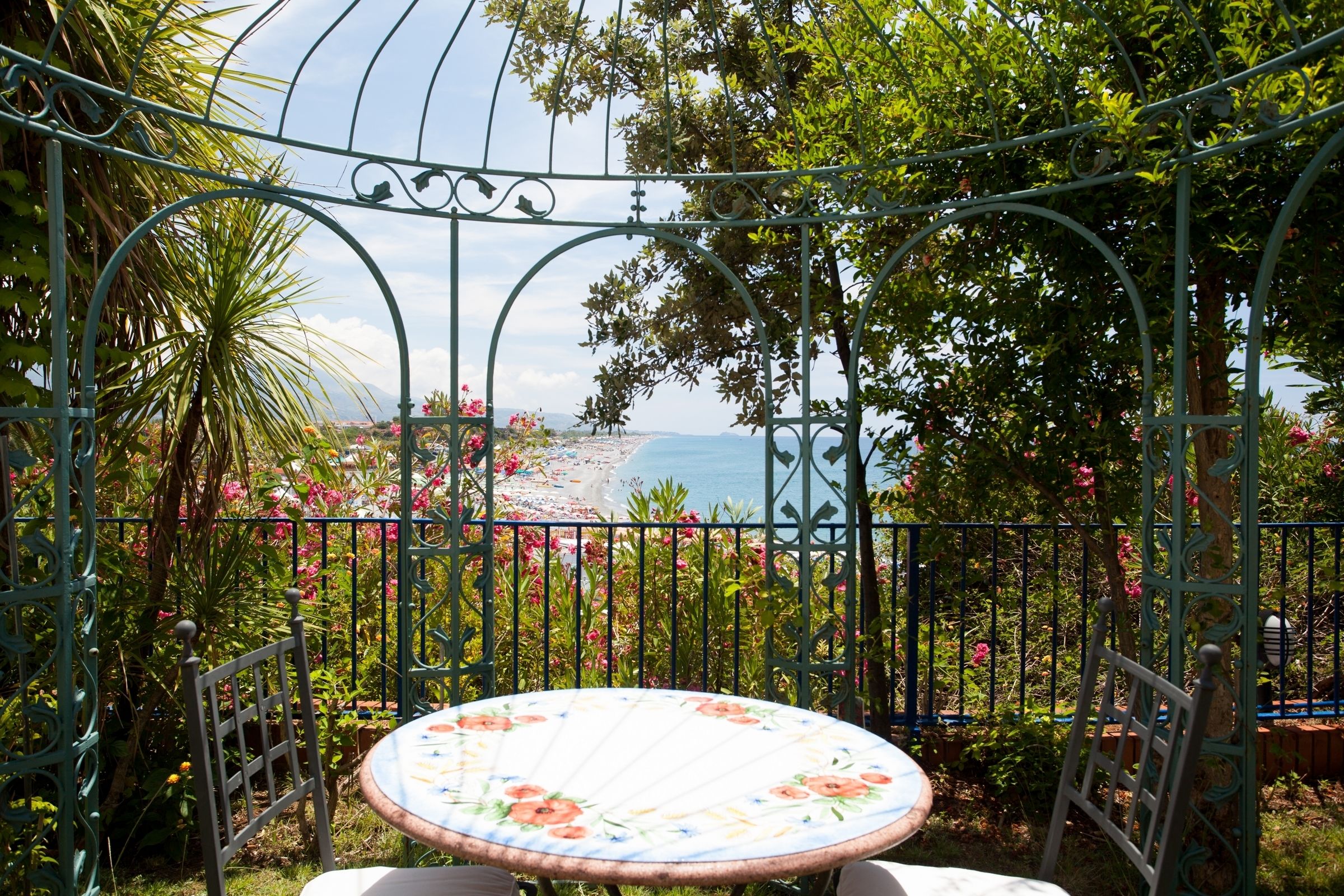 A garden with a metal fence, a table with a floral tablecloth, and a view of the beach.