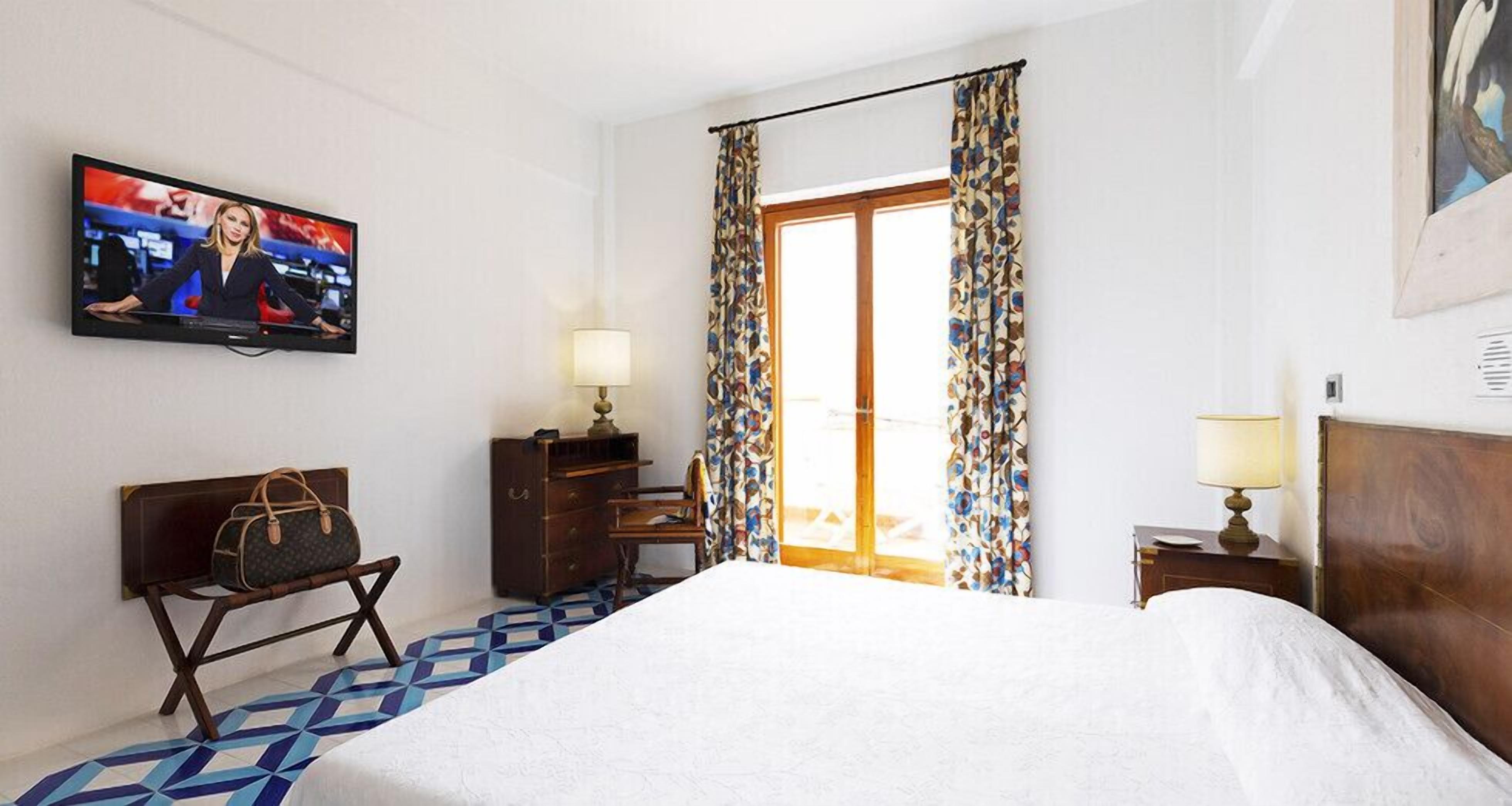 A hotel room with a bed, a desk with a lamp, a television on the wall, and a window with curtains.
