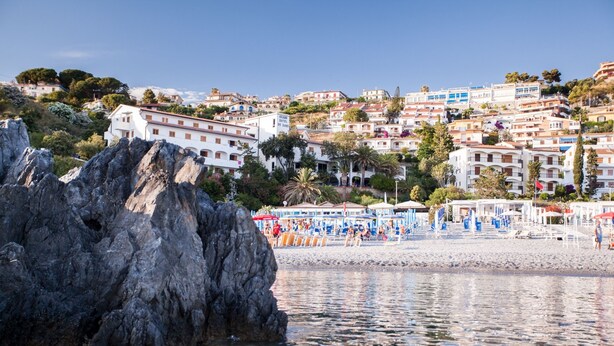 Une ville côtière avec des bâtiments construits sur une colline, un promontoire rocheux au premier plan et une plage parsemée de parasols et de personnes.