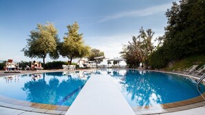 Une piscine à l’eau d’un bleu limpide, entourée de chaises longues et de parasols, avec des arbres et un ciel dégagé en arrière-plan.