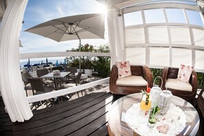 Une terrasse extérieure avec des meubles en osier, une table en verre avec des boissons, et une vue sur la mer.