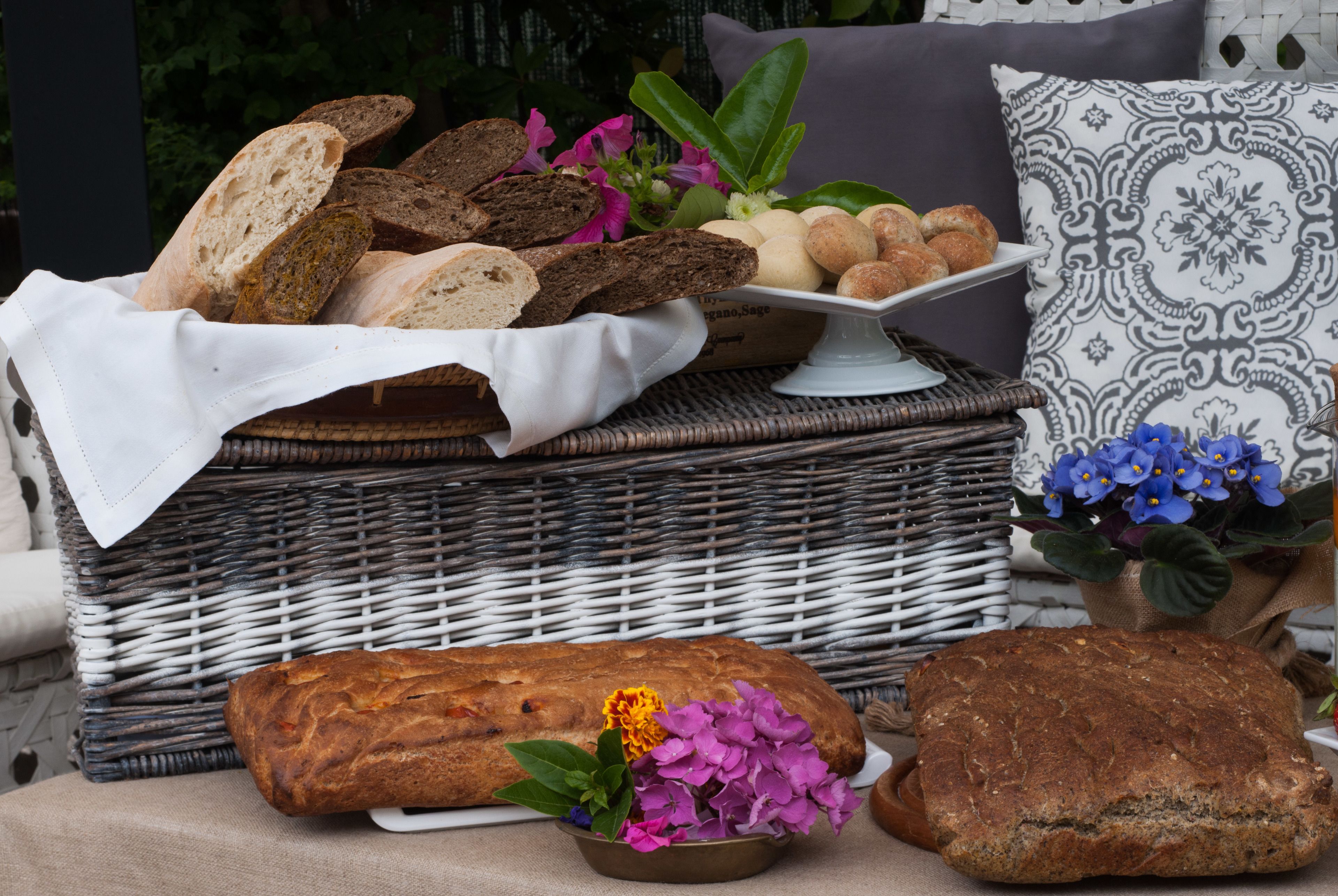 A wicker basket filled with various breads and pastries, accompanied by a loaf of bread and a small bowl of flowers.