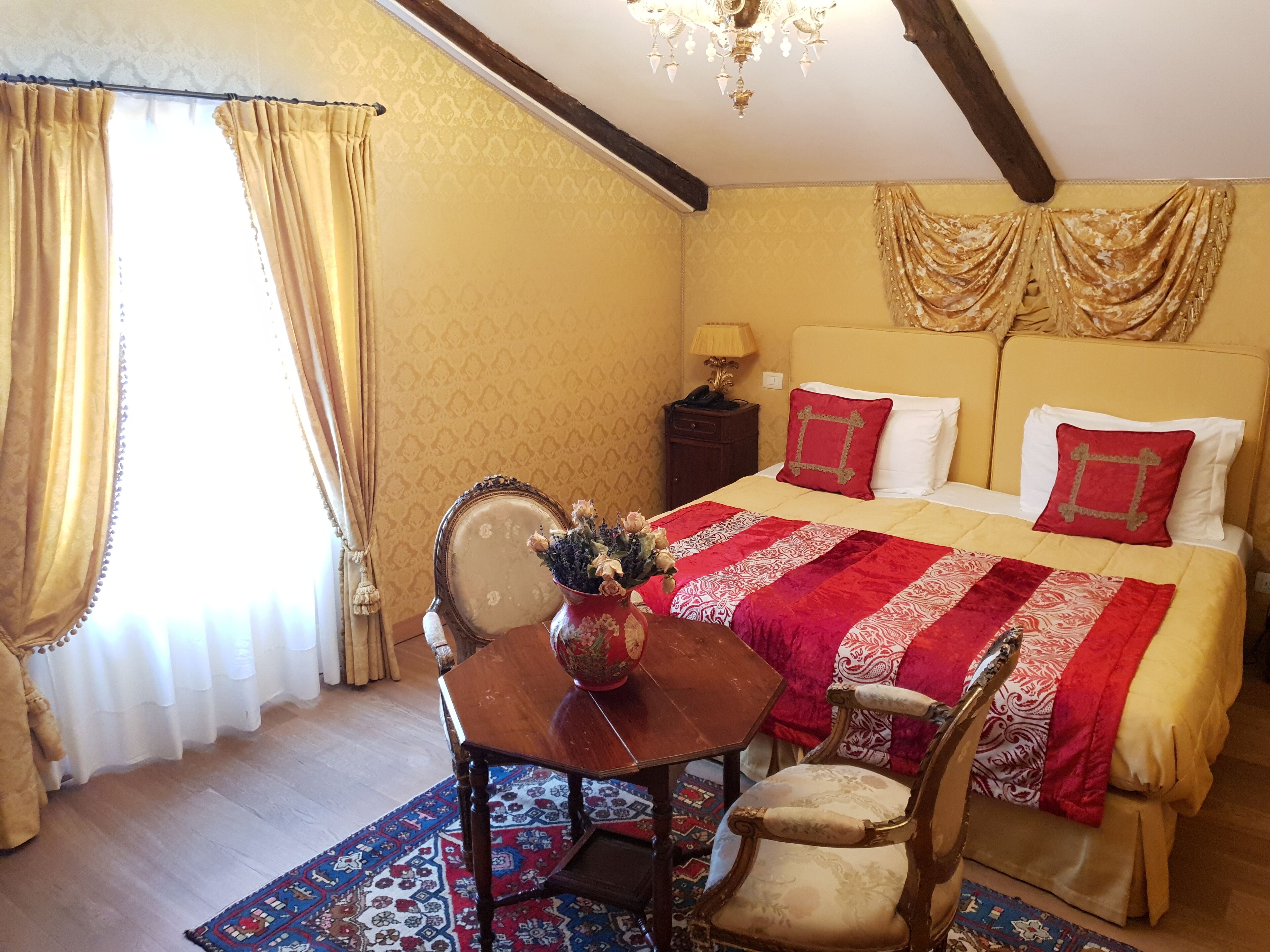 A room with a bed, a table with a vase of flowers, a chair, a lamp, and a patterned rug.