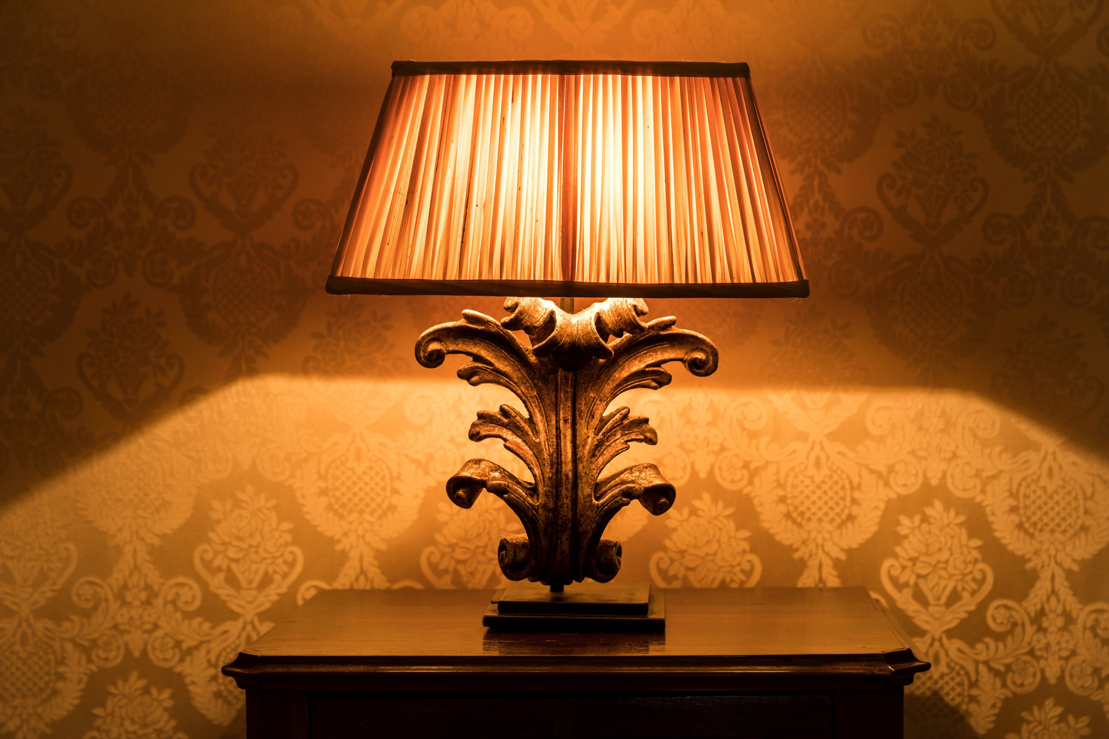Une lampe dotée d’un pied décoratif, posée sur une surface en bois, avec un papier peint à motifs en arrière-plan.