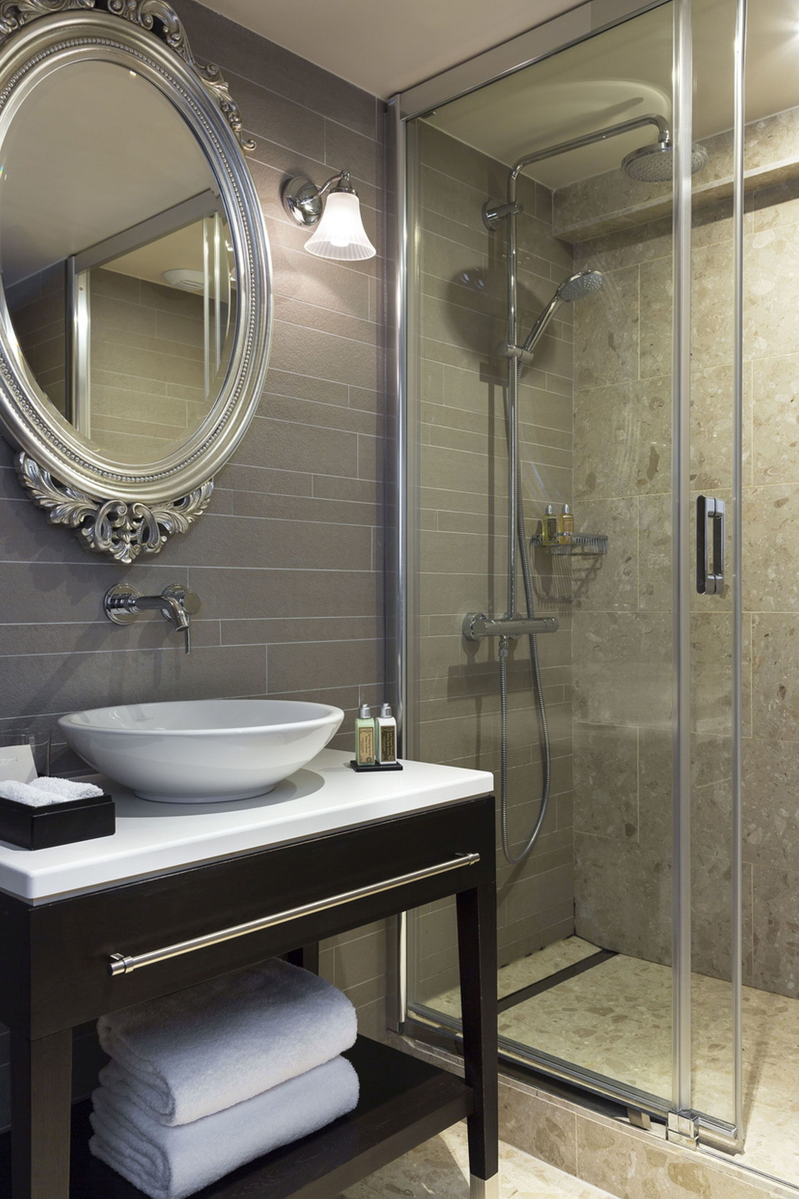 Un baño moderno con un espejo ovalado grande, un lavabo blanco tipo recipiente, un mueble de tocador de madera oscura y una zona de ducha con mampara de cristal.