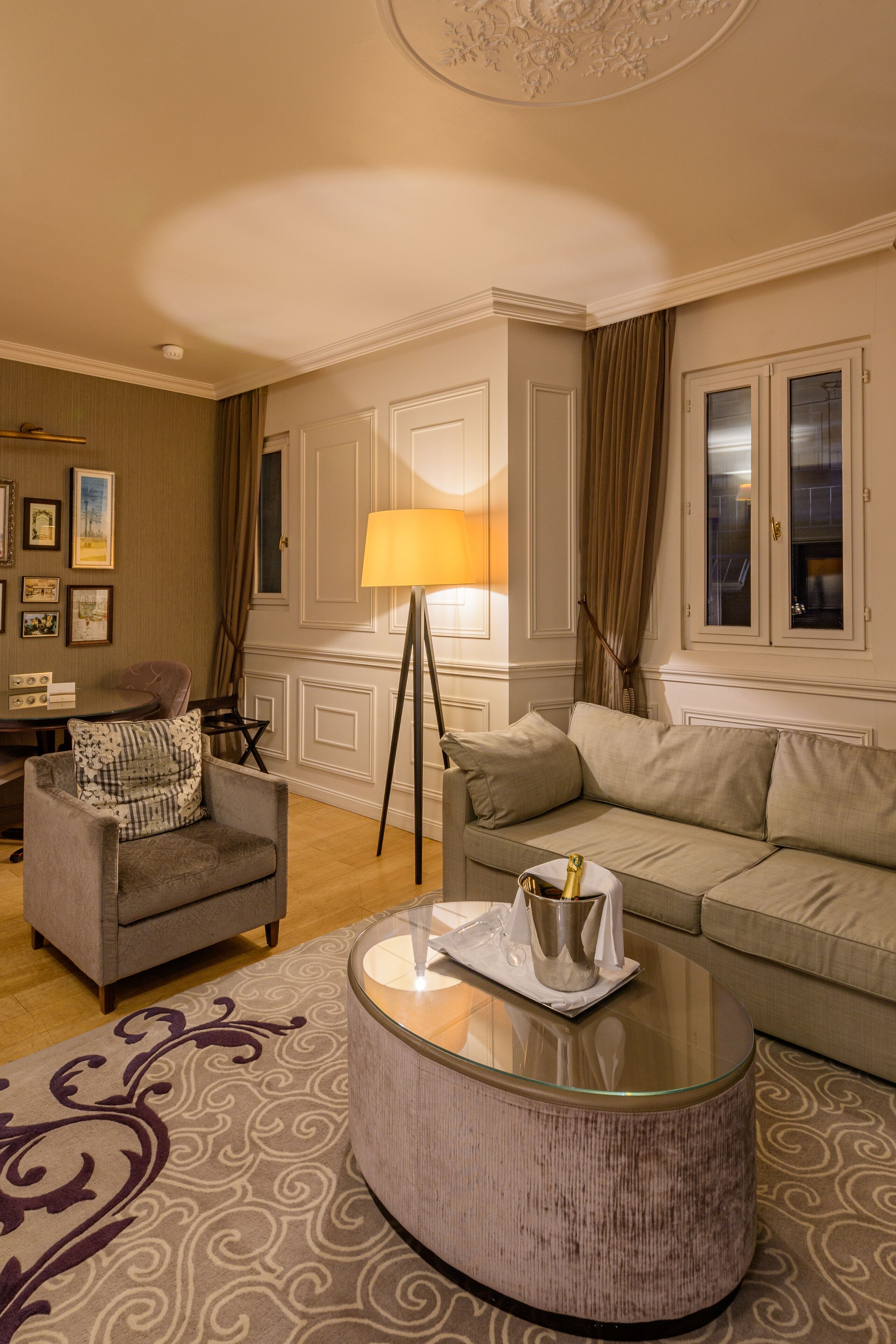 A well-lit living room with a round glass coffee table, a grey armchair with a patterned pillow, a beige sofa, and a large window with curtains.