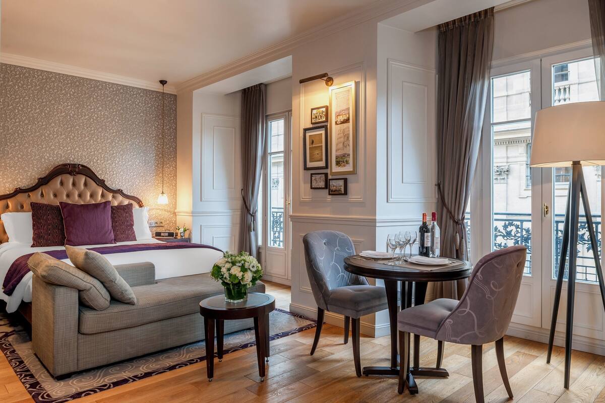 Ein Hotelzimmer mit einem Bett, einer Couch, einem Esstisch mit Stühlen, einer Blumenvase und Blick auf die Stadt durch große Fenster.