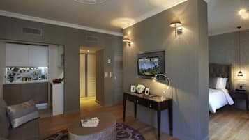 Ein modernes Hotelzimmer mit Holzboden, einem runden Couchtisch, einer Sitzecke, einem Bett mit weißer Bettwäsche, einem an der Wand montierten Fernseher und einem kleinen Schreibtisch mit Lampe.