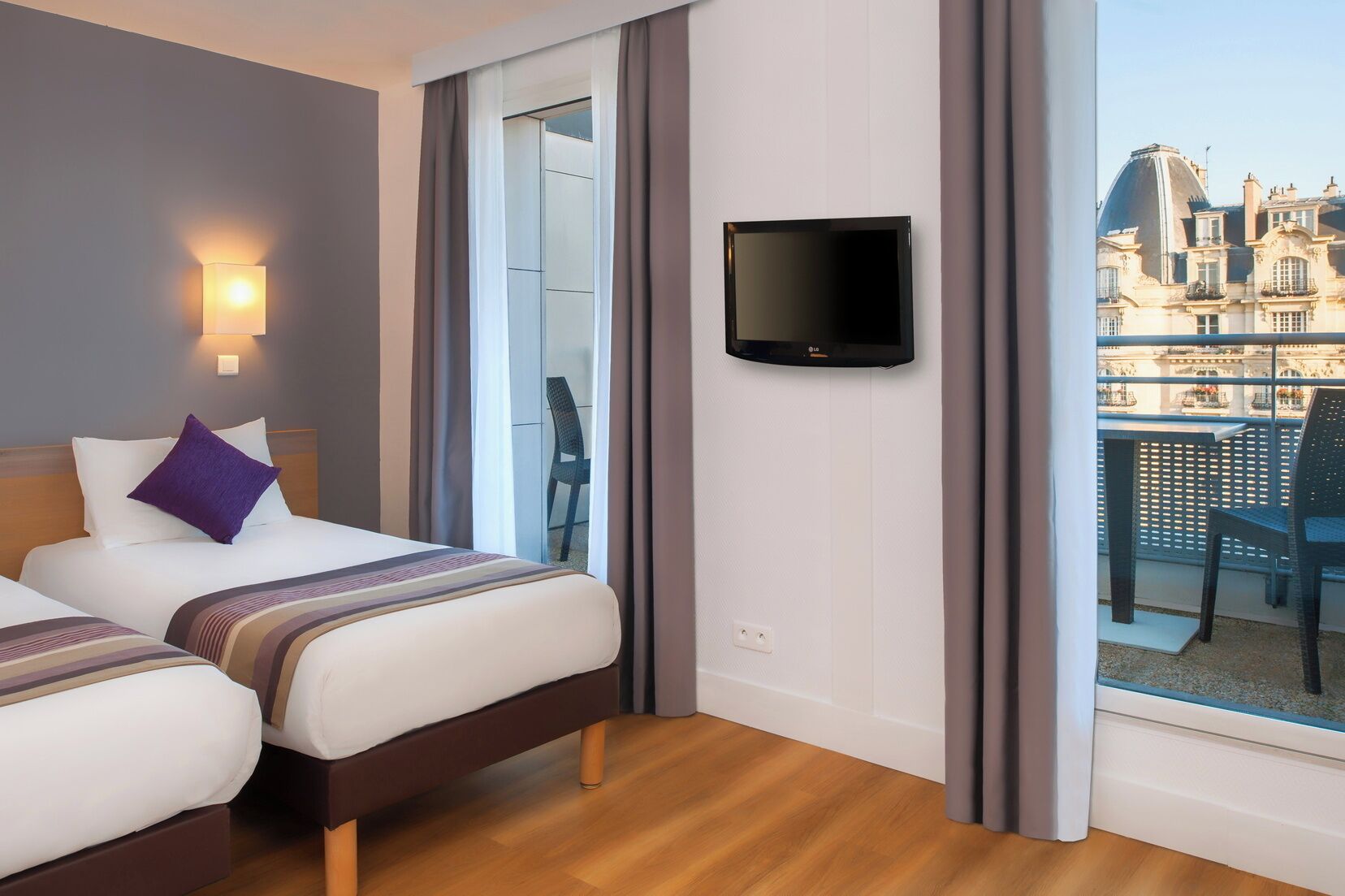 A hotel room with two beds, a television, and a balcony with a view of buildings.