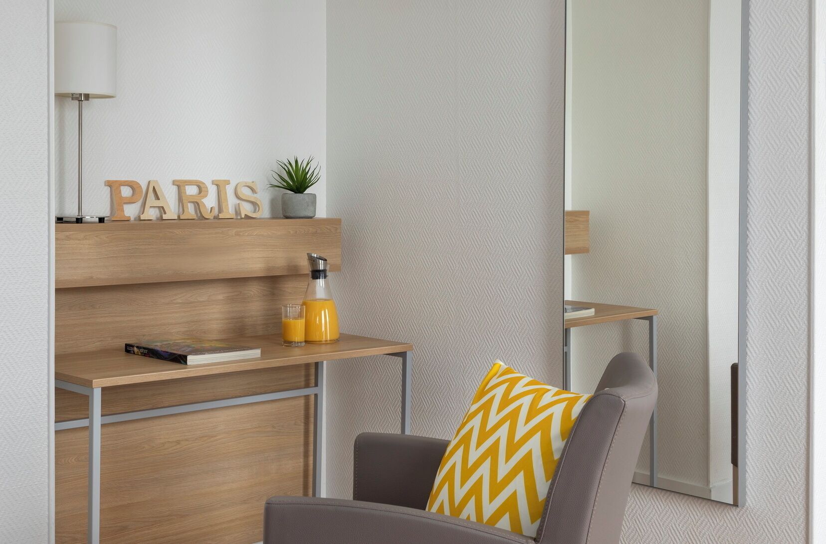 A modern room with a wooden desk, a chair with a patterned cushion, a mirror, and a wall-mounted shelf with a plant and a bottle.