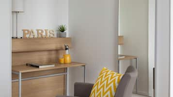 Una habitación moderna con un escritorio de madera, una silla con cojín estampado, un espejo y una estantería en la pared con una planta y una botella.