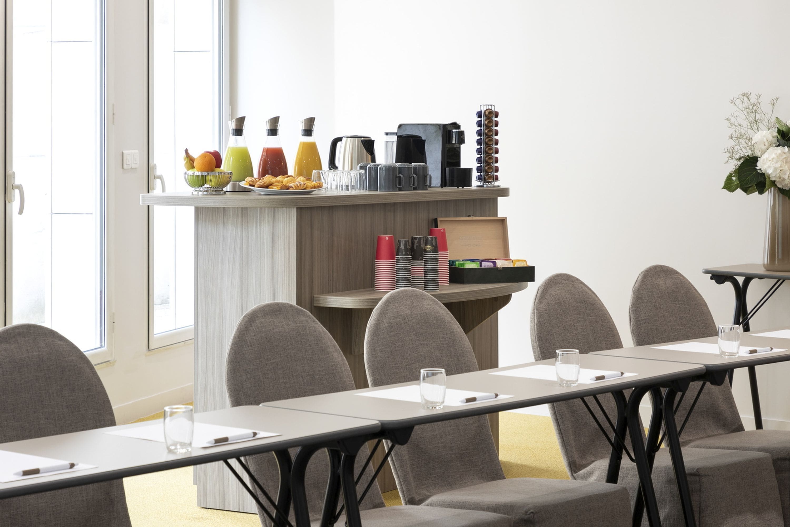 Une salle de conférence avec une longue table, des chaises grises, un coin café proposant diverses boissons et collations, et un vase de fleurs blanches posé sur une petite table d’appoint.