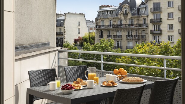 Un balcone con un tavolo apparecchiato per la colazione, con croissant, frutta e succo, affacciato su una città con edifici storici.