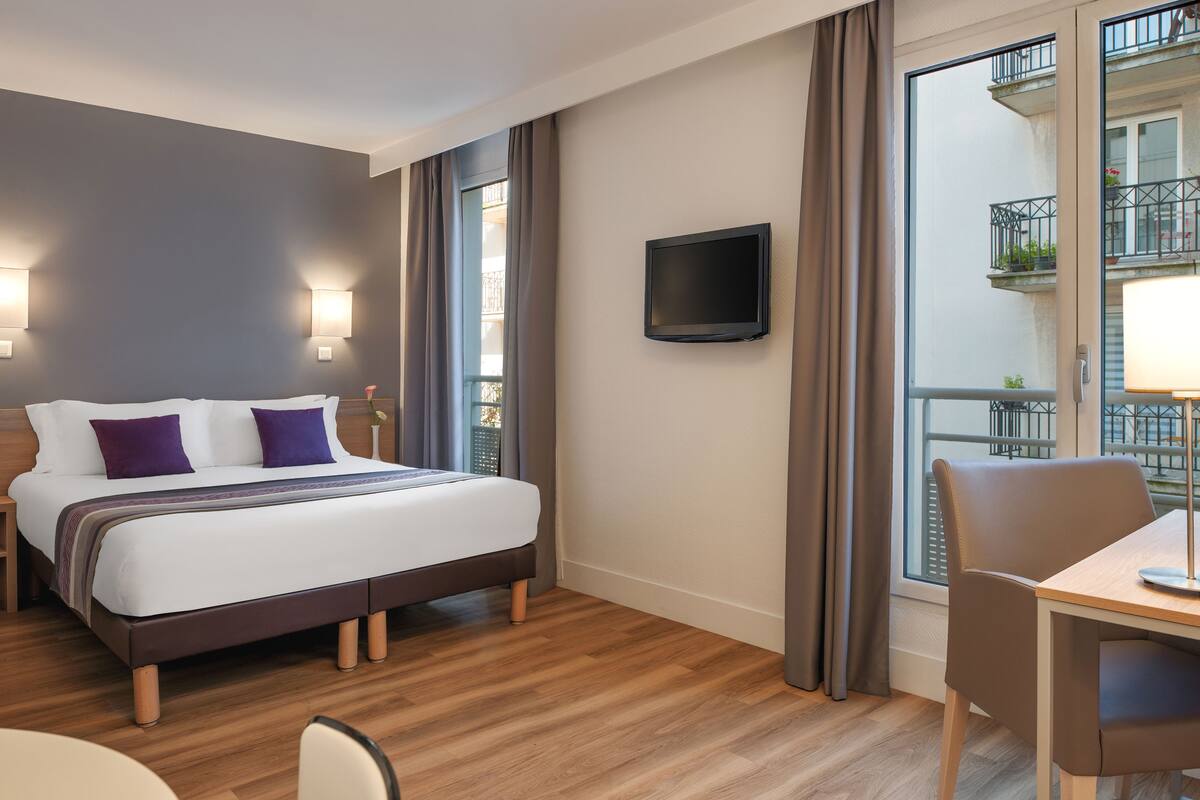 Habitación de hotel con cama, televisión, mesa de comedor y vista a un balcón con plantas.