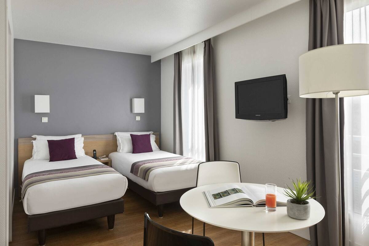 Habitación de hotel con dos camas, televisión, una mesa con un libro y una bebida, y una ventana con cortinas.