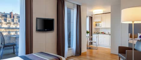 A hotel room with a bed, a TV, a view of the city, a kitchenette, and a dining area.