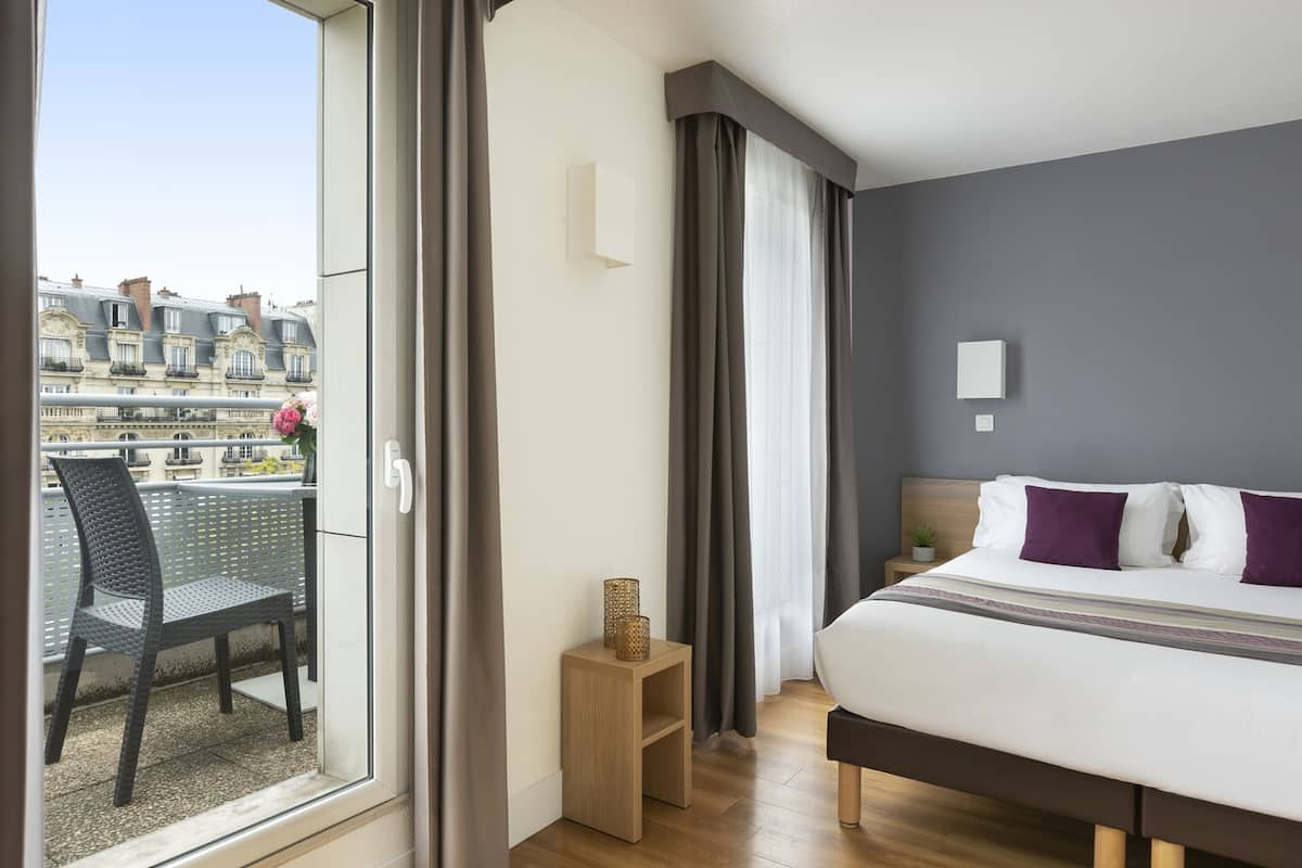 A hotel room with a balcony, a bed with white and purple bedding, a small wooden nightstand, and a view of buildings across the water.