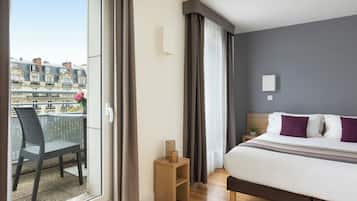 Habitación de hotel con balcón, cama, mesita de noche y vistas a edificios.